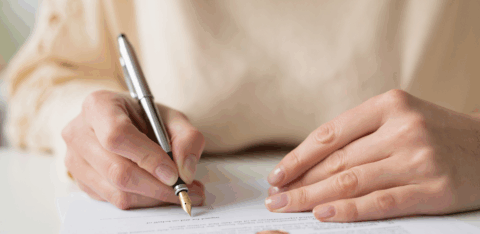Hands holding a pen and signing legal paperwork, with a wedding ring resting on the paper.