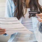 Woman holding a credit card in one hand and a bill in the other while reviewing finances.