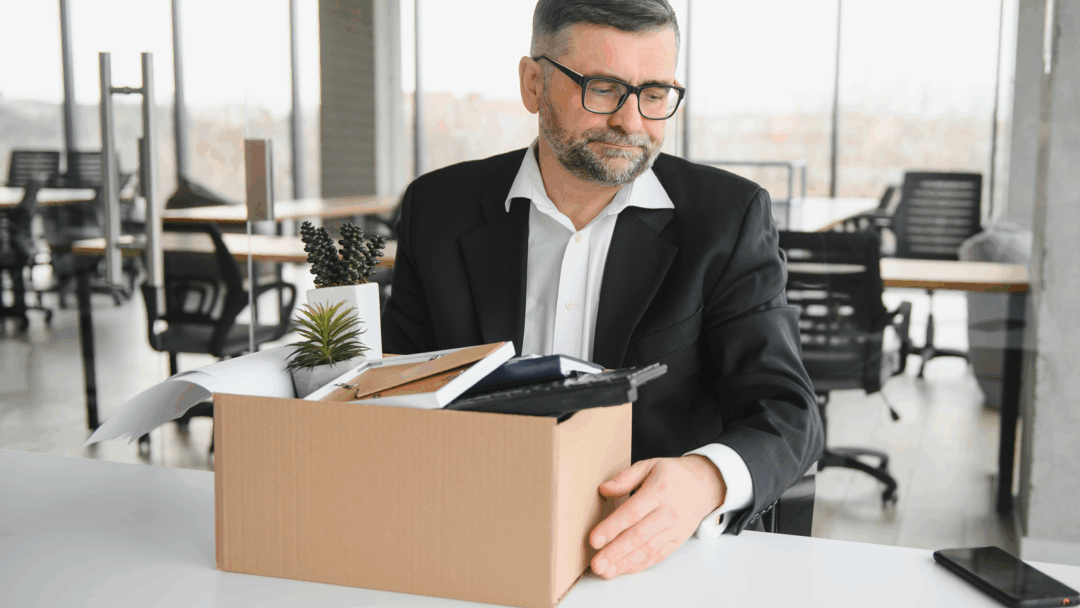 A man with glasses looking down sadly at a box of office items, suggesting he has been laid off or is leaving his job.