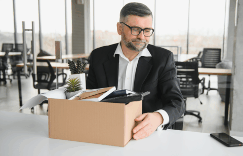 A man with glasses looking down sadly at a box of office items, suggesting he has been laid off or is leaving his job.
