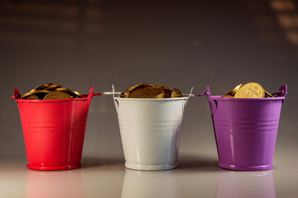 Metal buckets in red, white, and purple, each overflowing with gold coins against a neutral background.