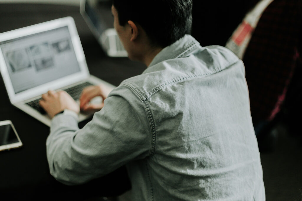Person in a denim shirt working on a laptop at a dark table, viewed from behind.