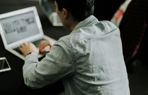 Person in a denim shirt working on a laptop at a dark table, viewed from behind.