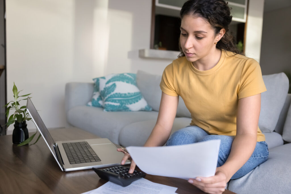 A woman sitting on a couch using a calculator and reviewing financial documents with a laptop open on the coffee table.