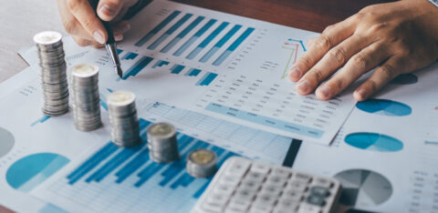 Person analyzing financial data with charts and stacked coins on a desk.