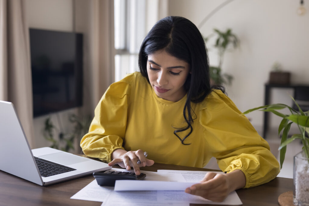 A woman in a yellow blouse calculating expenses with a calculator while reviewing documents at a table with a laptop.