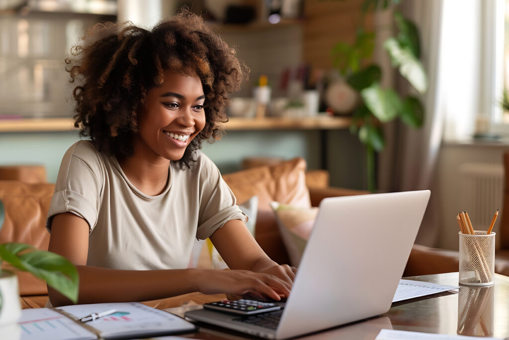 A smiling woman working on a laptop at home with charts and a calculator on the table.