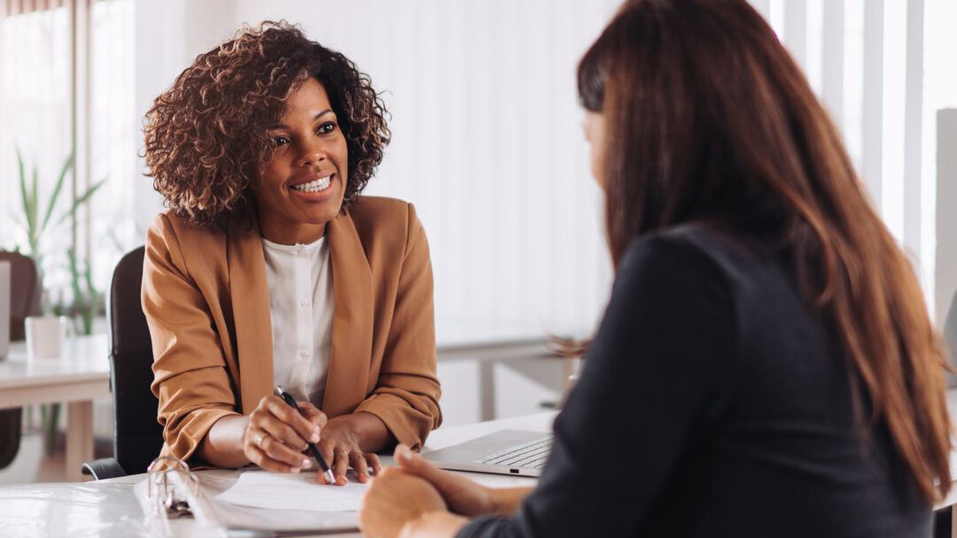 A professional woman in a tan blazer smiling while speaking with a client across a desk in a modern office setting.