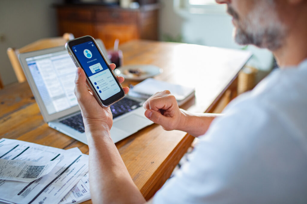 Person reviewing digital banking app on mobile phone with bills, receipts, and an open laptop on a wooden desk.