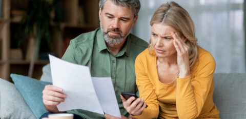 Two adults examine a document together at home, appearing worried as they check information on a phone.