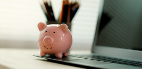 Small piggy bank placed on a laptop computer with blurred pencils in the background on a home office desk.