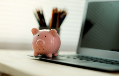Small piggy bank placed on a laptop computer with blurred pencils in the background on a home office desk.