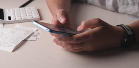 Person using a smartphone to review expenses at a desk with receipts and a calculator nearby.