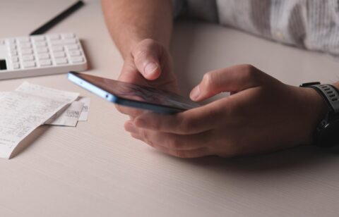 Person using a smartphone to review expenses at a desk with receipts and a calculator nearby.