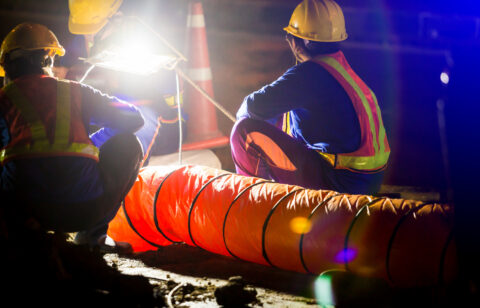 Road construction workers in safety vests and hard hats inspect equipment under bright work lights at night