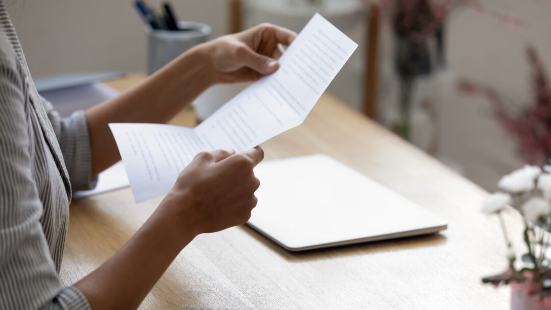 A person sits at a desk reading a printed document, with a closed laptop and notebook nearby.