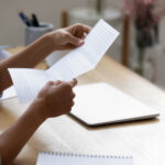 A person sits at a desk reading a printed document, with a closed laptop and notebook nearby.