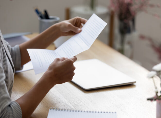 A person sits at a desk reading a printed document, with a closed laptop and notebook nearby.