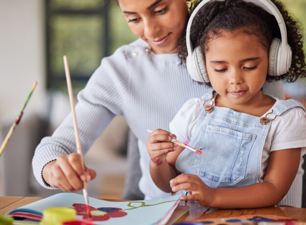 Mother and daughter painting together at home, with the child wearing headphones while creating artwork.