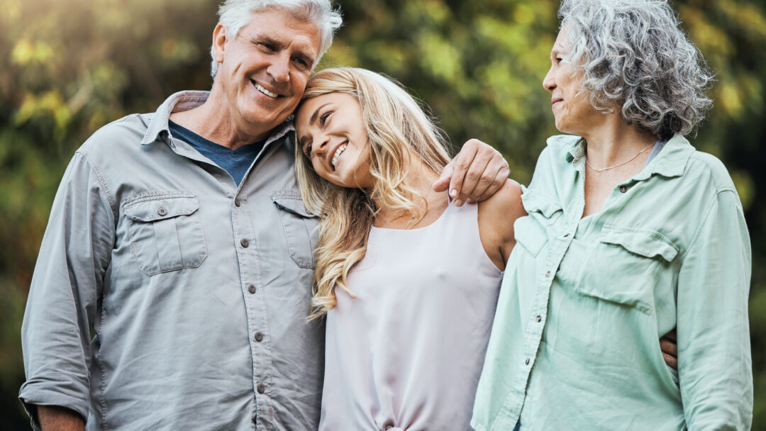 A family of three adults shares a warm moment outside, with the daughter resting her head on her father’s shoulder while her mother looks on.