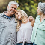 A family of three adults shares a warm moment outside, with the daughter resting her head on her father’s shoulder while her mother looks on.