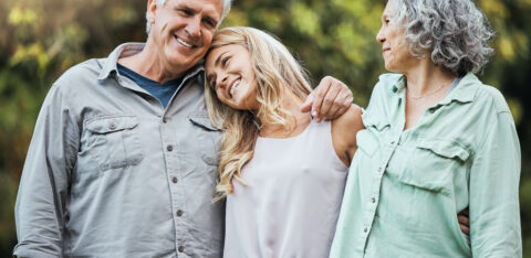 A family of three adults shares a warm moment outside, with the daughter resting her head on her father’s shoulder while her mother looks on.