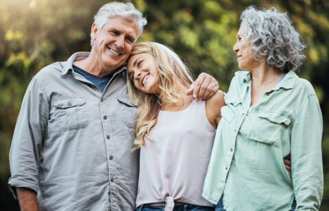 A family of three adults shares a warm moment outside, with the daughter resting her head on her father’s shoulder while her mother looks on.