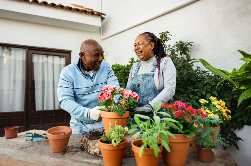 Older couple smiling while gardening together on a patio surrounded by colorful potted flowers and plants.