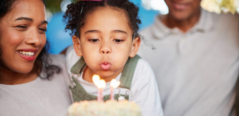 Child wearing a party hat blowing out birthday candles on a cake while parents smile nearby