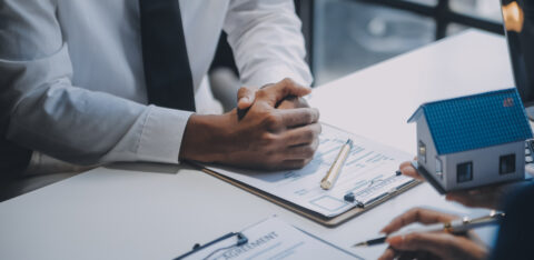 Close-up of two people at a desk with contract paperwork, a model house, and a pen