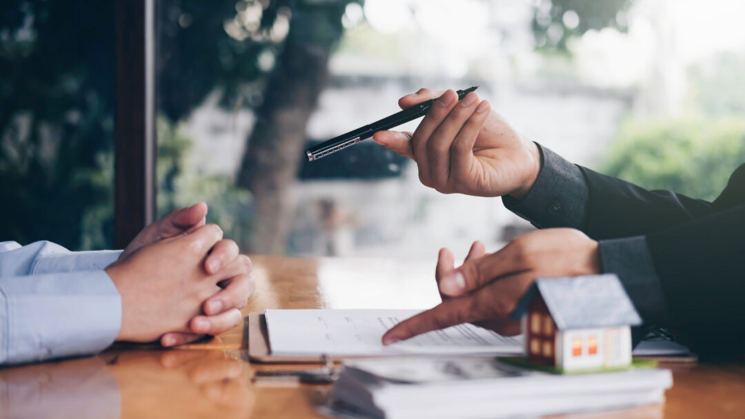 Financial professional discussing paperwork with a client at a desk with a small model house in the foreground.