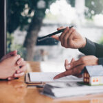 Financial professional discussing paperwork with a client at a desk with a small model house in the foreground.