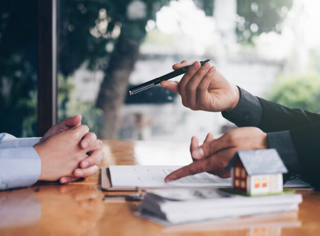 Financial professional discussing paperwork with a client at a desk with a small model house in the foreground.