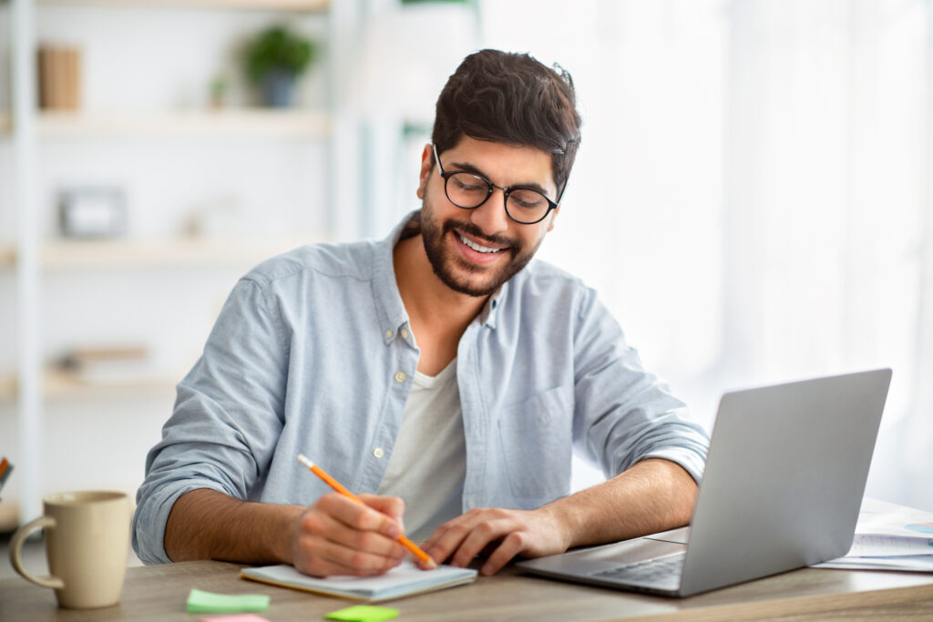 Happy man writing business report on laptop and taking notes, sitting at workplace at home interior
