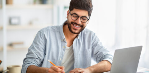 Happy man writing business report on laptop and taking notes, sitting at workplace at home interior
