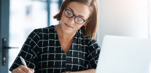 Professional woman writing in a notebook while working on a laptop in a modern office.