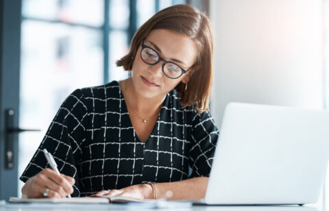 Professional woman writing in a notebook while working on a laptop in a modern office.