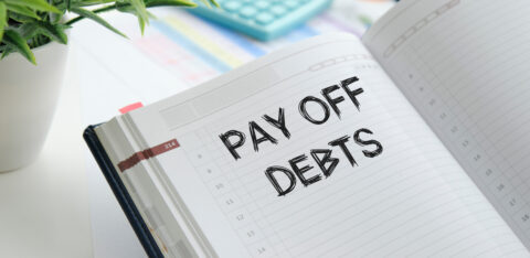 An open planner with the words "Pay Off Debts" written in bold, black font, surrounded by office supplies like a calculator, glasses, and a plant.