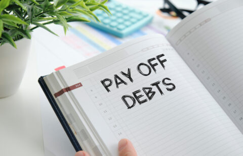 An open planner with the words "Pay Off Debts" written in bold, black font, surrounded by office supplies like a calculator, glasses, and a plant.