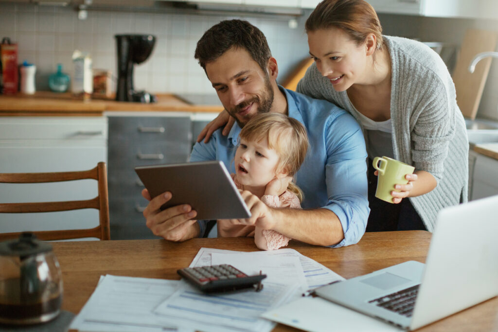 A happy family sitting at the kitchen table, reviewing financial documents on a tablet, with a laptop and calculator nearby.