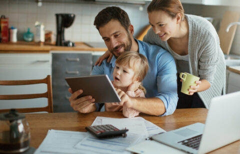 A happy family sitting at the kitchen table, reviewing financial documents on a tablet, with a laptop and calculator nearby.