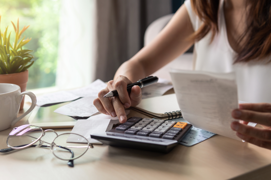 A woman calculating expenses at a desk, holding a receipt while using a calculator, with financial documents and a notebook in front of her.