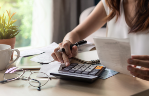 A woman calculating expenses at a desk, holding a receipt while using a calculator, with financial documents and a notebook in front of her.
