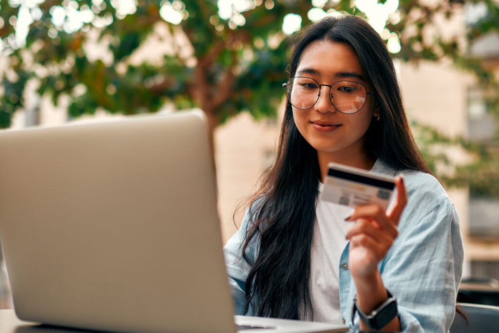 A young woman wearing glasses and a smartwatch holds a credit card while using her laptop outdoors.