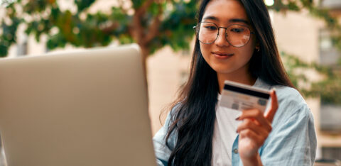 A young woman wearing glasses and a smartwatch holds a credit card while using her laptop outdoors.