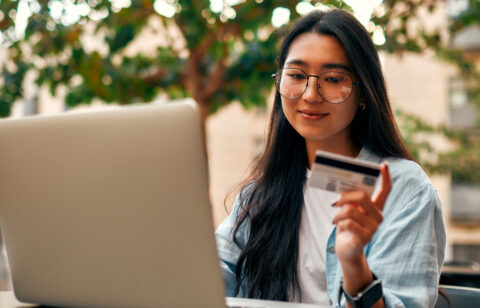 A young woman wearing glasses and a smartwatch holds a credit card while using her laptop outdoors.