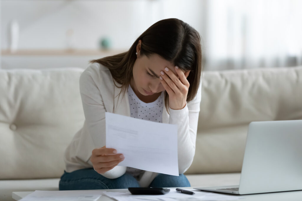A worried woman sitting on a couch, holding a bill in one hand and resting her head on the other, surrounded by financial documents and a laptop.