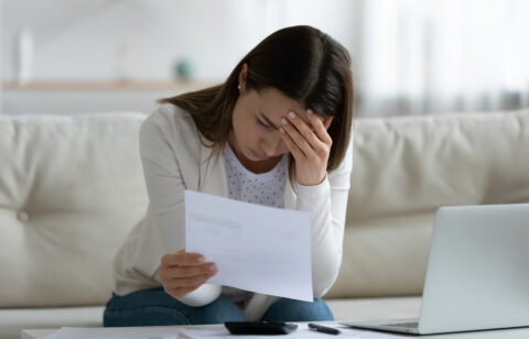 A worried woman sitting on a couch, holding a bill in one hand and resting her head on the other, surrounded by financial documents and a laptop.
