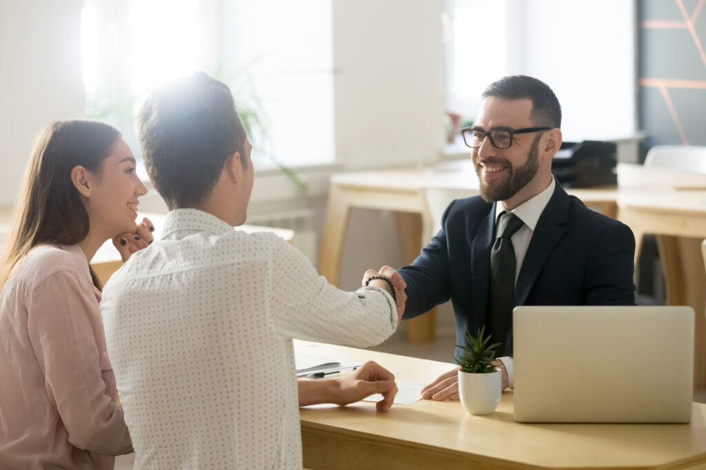 A smiling financial advisor in a suit shakes hands with a male client, while a female client watches with a smile in a modern office setting.