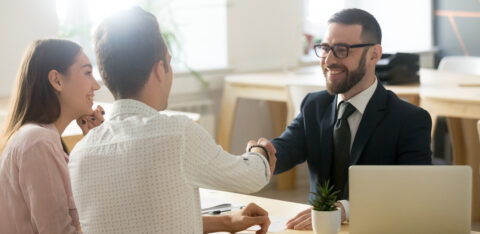 A smiling financial advisor in a suit shakes hands with a male client, while a female client watches with a smile in a modern office setting.
